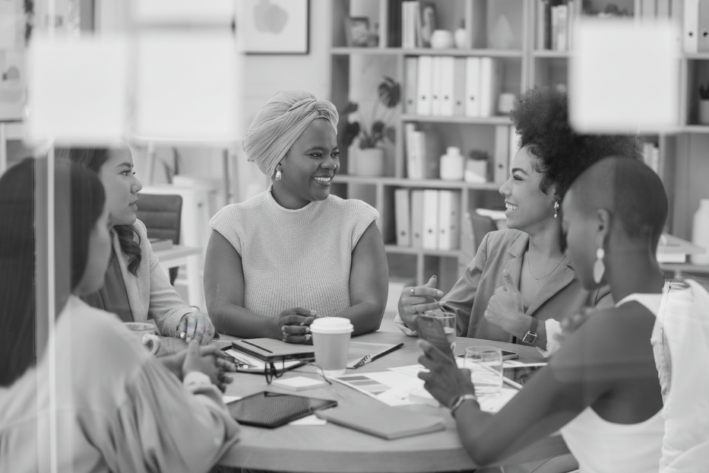 A diverse group of women sitting around a table in a bright office, smiling and engaged in a collaborative discussion with notebooks, coffee, and planning materials.