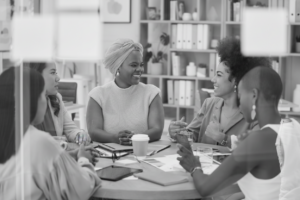 A diverse group of women sitting around a table in a bright office, smiling and engaged in a collaborative discussion with notebooks, coffee, and planning materials.