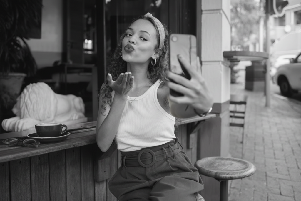 A woman sitting at an outdoor café takes a selfie with her phone while blowing a kiss, with a coffee cup on the counter beside her.