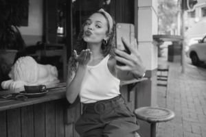 A woman sitting at an outdoor café takes a selfie with her phone while blowing a kiss, with a coffee cup on the counter beside her.