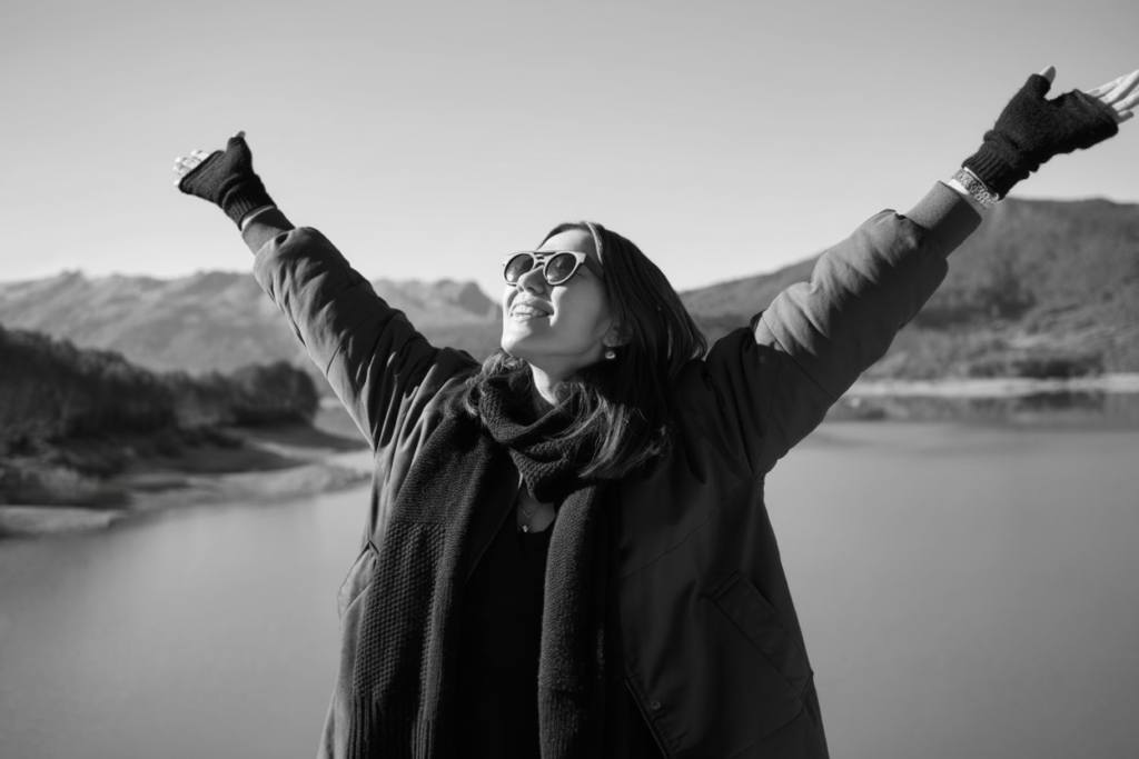 A woman standing outside with a lake view and hills in the background with her hands up towards the sky. She is wearing sunglasses, a green jacket, and gloves.