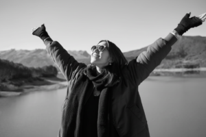 A woman standing outside with a lake view and hills in the background with her hands up towards the sky. She is wearing sunglasses, a green jacket, and gloves.