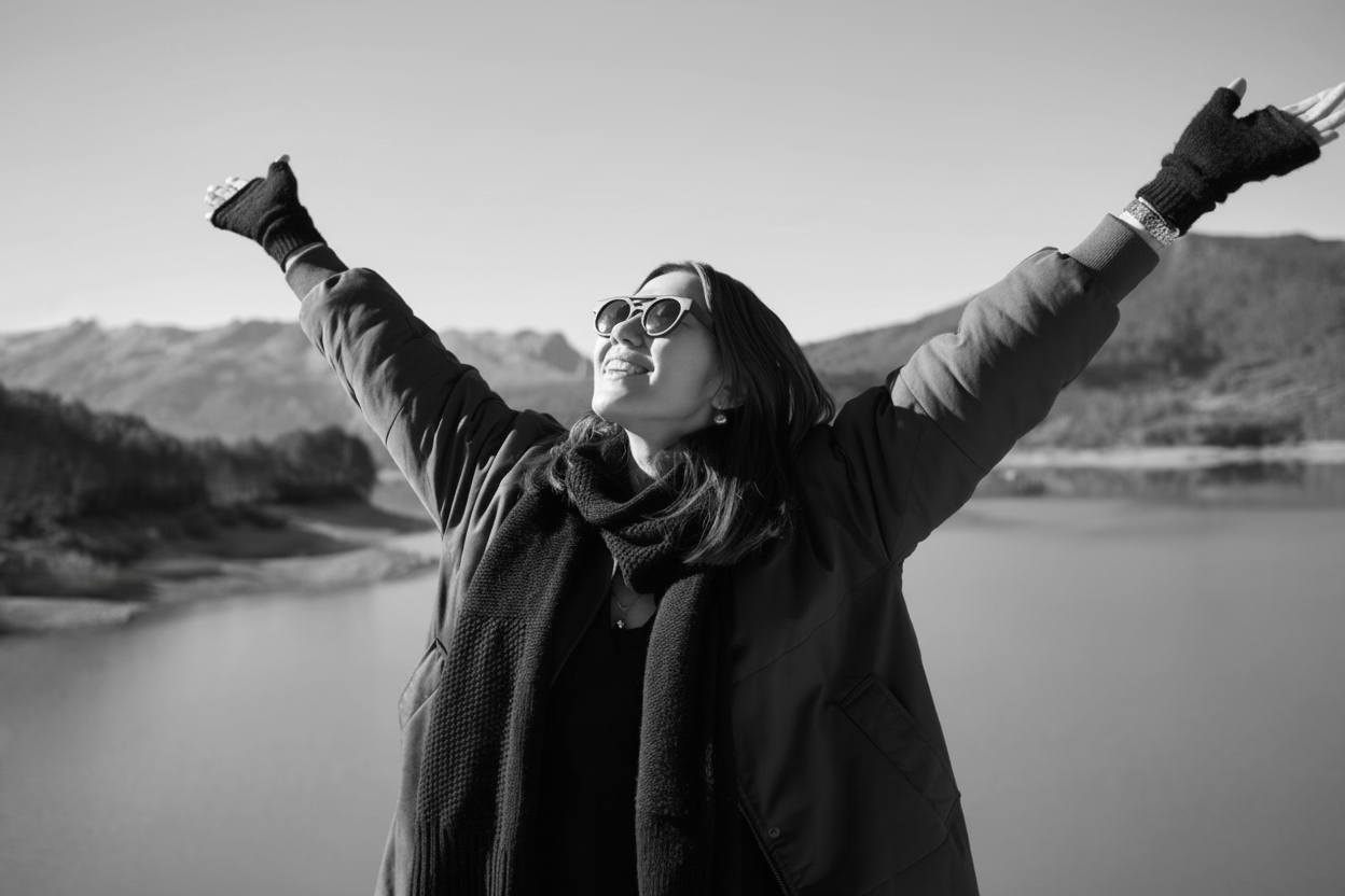 A woman standing outside with a lake view and hills in the background with her hands up towards the sky. She is wearing sunglasses, a green jacket, and gloves.